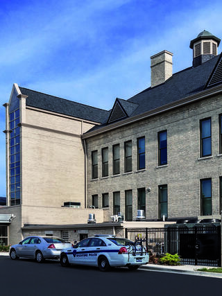A modern building features brick architecture with a flat roof. Two police vehicles are parked nearby, set against a clear blue sky.