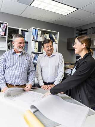 Three professionals engage in a discussion while reviewing plans, displaying smiles and a collaborative atmosphere in an office setting.