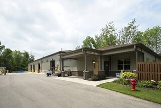 A modern, single-story industrial building with a parking area, surrounded by greenery and a fire hydrant in a peaceful setting.
