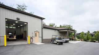 A modern, gray metal building features an open bay with equipment inside and parked golf carts outside, surrounded by greenery.