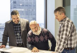 Three men are discussing architectural plans while standing around a table, with a cityscape visible through large windows in the background.