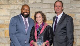 Three individuals smile together, with a woman holding an award, set against a stone wall background.