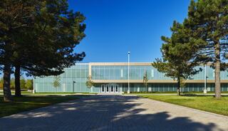A modern, glass-fronted building flanked by trees, with a clear blue sky and a paved walkway leading to the entrance.