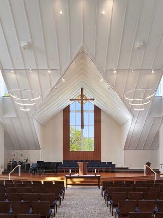 A bright, spacious interior featuring a tall wooden cross, large windows, and rows of seats, designed for gatherings and worship.