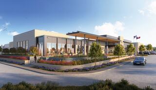 A modern building features large glass windows, landscaped greenery, and flags, set against a bright blue sky, with a welcoming entrance.