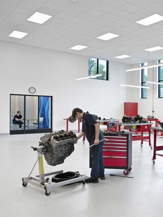 A mechanic works on an engine in a bright, spacious workshop with red tool carts and a person seated in the background.