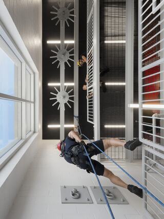A firefighter rappels down a high space, wearing harnesses, while others observe from above, amidst modern architecture and safety features.
