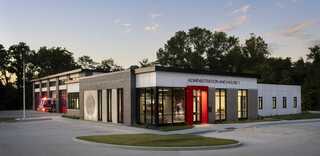 A modern building featuring a sleek design, large windows, and red accents, surrounded by greenery and parking space.