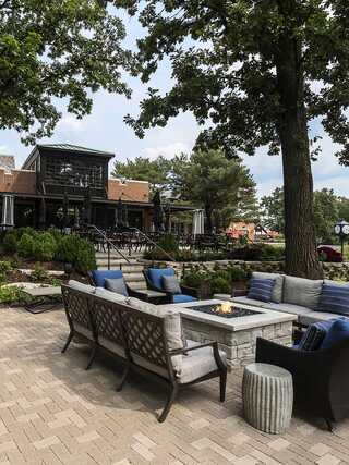 A cozy outdoor seating area features a fire pit surrounded by lush greenery, leading to an inviting building in the background.