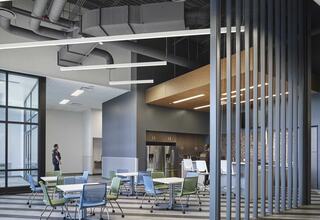 A modern workspace features sleek tables and chairs, large windows, and a stylish kitchen area, creating an open and inviting atmosphere.