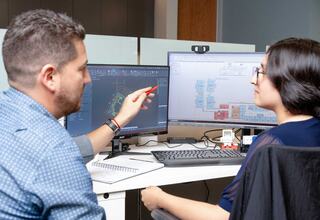 Two professionals collaborate at work, discussing design plans displayed on computer screens, with one actively pointing out details.
