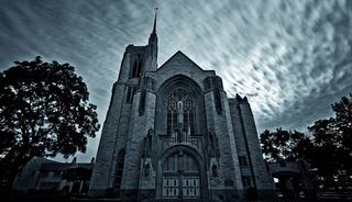A grand, gothic-style church stands against a moody, cloud-filled sky, surrounded by dark trees, creating an eerie yet striking atmosphere.