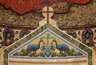 A colorful mosaic features two blue peacocks beside a vase, adorned with a cross above them, set against a rich, patterned background.