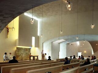 A modern church interior featuring wooden pews, soft lighting, and a minimalist altar space, with a small congregation in attendance.