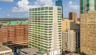 A modern high-rise building with green-tinted windows stands amidst a mix of contemporary and historic architecture under a blue sky.