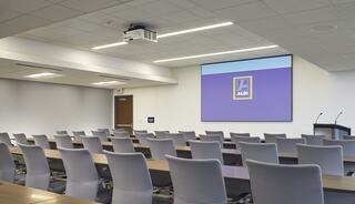 A modern conference room with rows of tables and chairs, featuring a projector screen displaying the Aldi logo on a purple background.