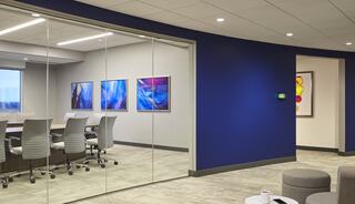 A modern office space features a glass-walled conference room with sleek chairs and colorful artwork, set against a vibrant blue wall.