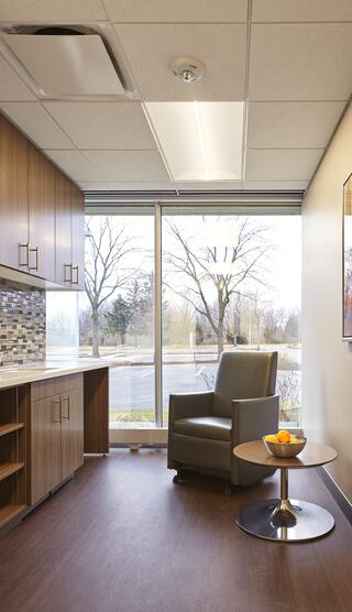 A modern, cozy room features a gray armchair beside a round table with fruit, large windows offering a view of trees and a parking lot.