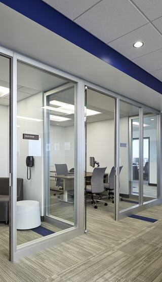 A modern office space featuring glass-walled rooms, desk chairs, and a phone. The design is sleek and professional with neutral tones.