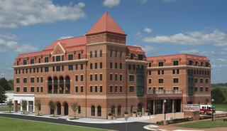 A large, modern brick building features a mix of architectural styles, with a red-tiled roof and multiple levels. Lush greenery surrounds it.