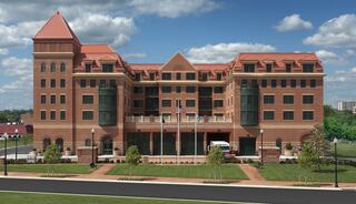 A modern, multi-story brick building features a distinct roof design and large windows, surrounded by landscaped greenery and a paved pathway.