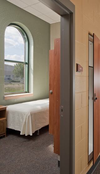 A serene room with a bed, a large arched window, and natural light. Wooden partitions and a simple, clean design create a restful ambiance.
