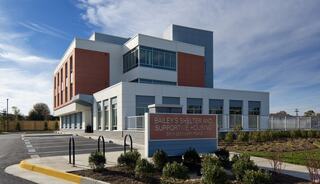 A modern, multi-story building with a mix of orange and white panels, featuring a sign for Bailey's Shelter and Supportive Housing. Garden space enhances the exterior.