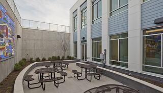A modern outdoor space features several circular picnic tables surrounded by greenery and a colorful mural on the adjacent wall.