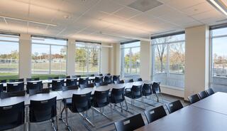 A bright, modern classroom features large windows, several tables, and black chairs, overlooking an outdoor view with trees and greenery.