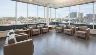 A modern waiting area features several brown chairs arranged neatly, large windows offering a view of an urban landscape. Bright and inviting space.