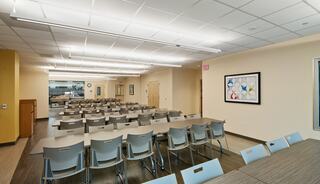 A spacious room with rows of tables and chairs, brightly lit, featuring a kitchenette and a decorative framed artwork on the wall.