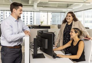 Three professionals engage in a lively conversation at a modern office space, with one seated at a computer while the others stand nearby.