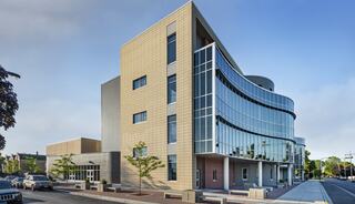 A modern building features a mix of glass and textured surfaces, with a curved facade and landscaped surroundings, creating a sleek urban aesthetic.