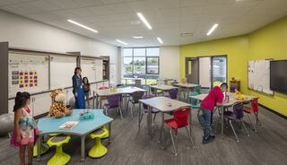 A bright classroom features colorful tables and chairs, playful decor, and engaged children interacting with a teacher.