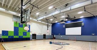 A spacious gym features basketball hoops, colorful climbing walls, and a projector screen, with wooden flooring and bright overhead lighting.