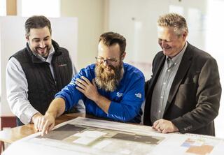Three men collaborate and discuss a large blueprint, engaging cheerfully as they point at different sections of the plans.