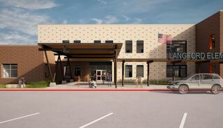 A modern school entrance features a large overhang, an American flag, and a welcoming design, surrounded by a parking area.