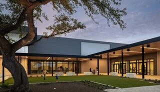 A modern building features large glass windows, outdoor seating, and a landscaped area with children playing under a mature tree at dusk.