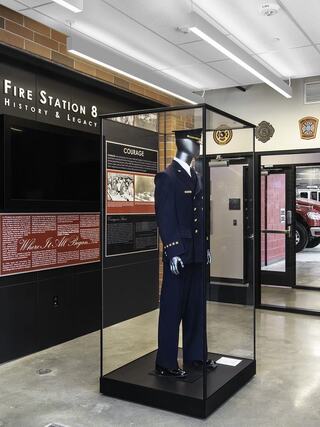 A display case showcases a firefighter's uniform, surrounded by informative panels about the history and legacy of Fire Station 8.