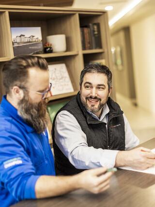 Two men engage in a friendly conversation at a table, with one taking notes. A relaxed office environment surrounds them.