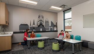A bright classroom features children working at tables, accompanied by adults. A mural of city buildings adorns one wall.