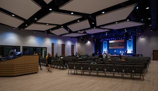 A spacious auditorium features rows of empty chairs, a stage with lights, and a modern design, suggesting preparation for an event.