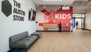 A welcoming lobby features a gray bench, a vibrant "KIDS" sign, and a reception area with kiosks for check-in, creating a family-friendly atmosphere.
