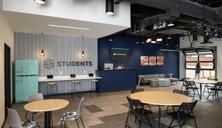 A modern student lounge features a welcoming reception area, blue walls, light wood accents, a vintage fridge, and communal tables for collaboration.