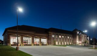 A modern fire station illuminated at night, showcasing large glass doors and a welcoming facade amidst a well-maintained parking lot.