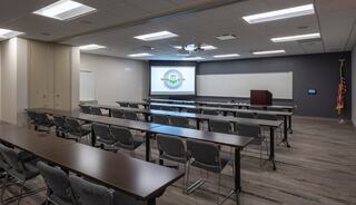 A spacious conference room with rows of tables and chairs, featuring a screen and podium at the front, ideal for meetings and presentations.
