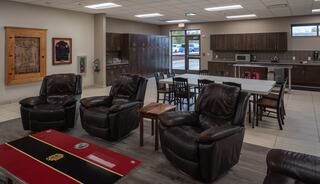 A cozy lounge features leather recliners, a dining area, a kitchen, and wooden cabinets, illuminated by natural light from large windows.
