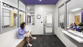 A woman sits on a bench, looking at her phone, while another person is engaged in a conversation behind glass in a modern, bright space.