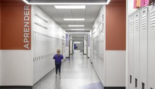 A school hallway features lockers on both sides, with soft lighting and a child in purple running toward a classroom. The word "APRENDER" is visible.