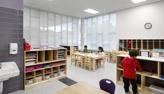 A bright, modern classroom features children engaged in activities around wooden tables, with organized shelves and large windows allowing natural light.